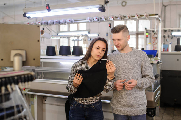Nahaufnahme Foto einer jungen Frau und eines jungen Mannes, die ein gestricktes Kleidungsstück betrachten und in der Nähe einer industriellen Strickmaschine mit schwarzen Kegeln diskutieren - Foto, Bild