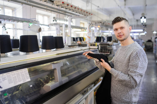 un giovane uomo che analizza un pezzo di stoffa lavorato a maglia vicino a macchine industriali a maglia con filo nero in coni  - Foto, immagini