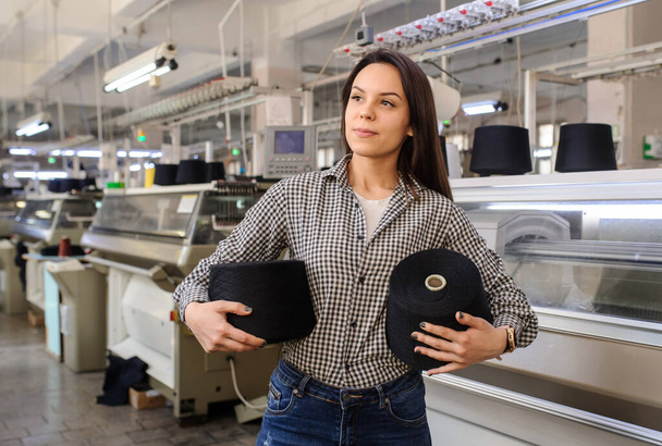 close up photo of a young woman with black threads in cones in her hands near industrial knitting machines - Photo, Image