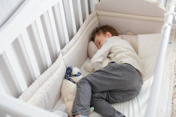 top view of a blond seven years boy sleeping in his sister white wooden cot in white shirt and grey trousers with toys in his arms - Photo, Image