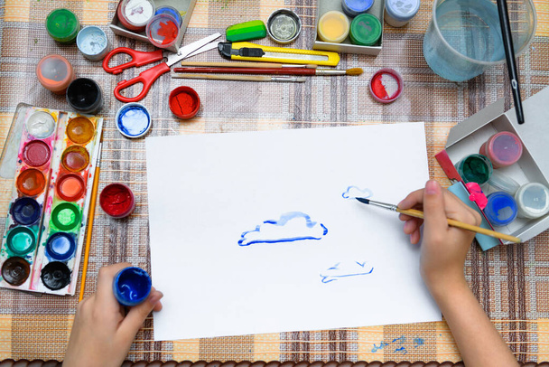 Beyaz bir kağıda suluboya mavi bulutlar çizen bir kız evde sanatsal bir eser yaratıyor. - Fotoğraf, Görsel