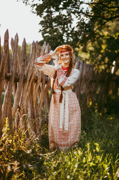 A beautiful Slavic girl with long blonde hair and brown eyes in a white and red embroidered suit stands by a wooden fence.Traditional clothing of the Ukrainian region.summer day - Foto, imagen