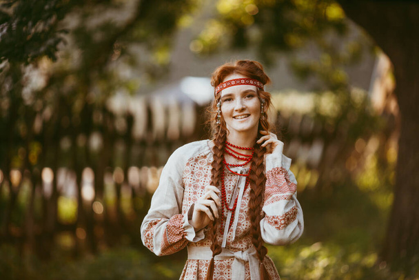 A beautiful Slavic girl with long blonde hair and brown eyes in a white and red embroidered suit stands by a wooden fence.Traditional clothing of the Ukrainian region.summer day - Fotoğraf, Görsel