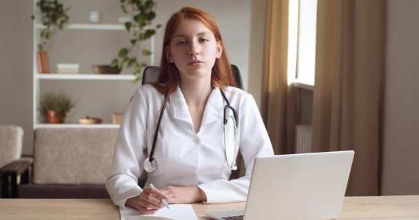 Молодая задумчивая женщина врач носит медицинскую форму, сидит в клинике с ноутбуком, смотрит в окно думает, поворачивает взгляд к камере, улыбаясь дружелюбно держа ручку в руках. Портрет медсестры - Кадры, видео
