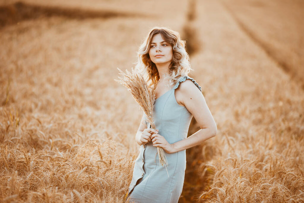 A young girl with long curly hair and a light blue dress , standing in a wheat field, posing for the camera, against the background of the sunset - Foto, Imagen