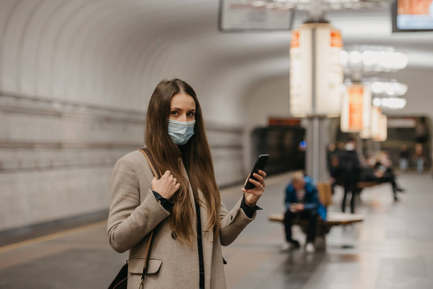 Жінка в масці обличчя, щоб уникнути поширення коронавірусу, виставляє смартфон на станції метро. Дівчина в масці для хірургічного обличчя проти COVID-19 чекає поїзд на платформі метро.. - Фото, зображення