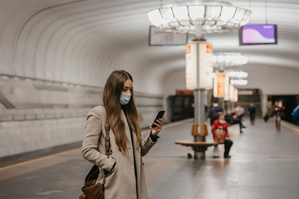 Een vrouw met een gezichtsmasker om de verspreiding van het coronavirus te voorkomen houdt een smartphone vast bij een metrostation. Een meisje met een chirurgisch masker op het gezicht tegen COVID-19 wacht op een trein op een metroperron - Foto, afbeelding