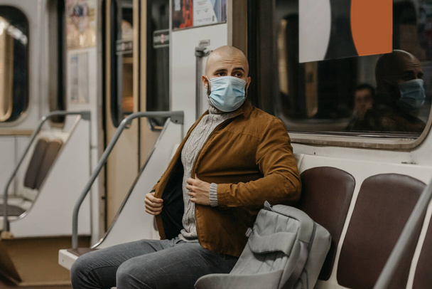 Людина з бородою в медичній масці на обличчі, щоб уникнути поширення коронавірусу їде в машині метро. Лиса людина в хірургічній масці обличчя проти COVID-19 знаходиться в поїзді метро.. - Фото, зображення