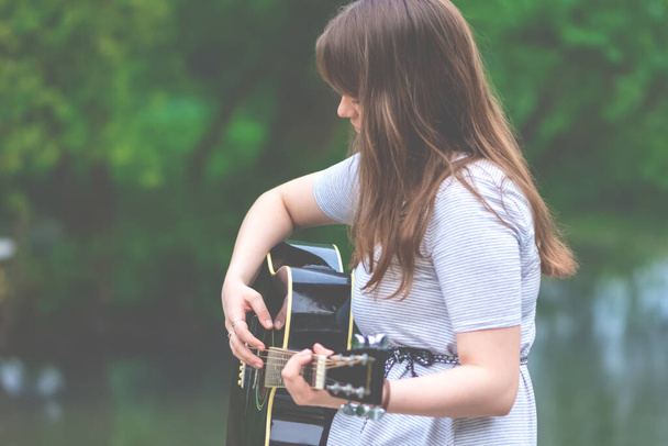 Ładna nastolatka gra na gitarze siedząc na kamiennym moście. Młoda kobieta gitarzysta odkryty w naturze - Zdjęcie, obraz