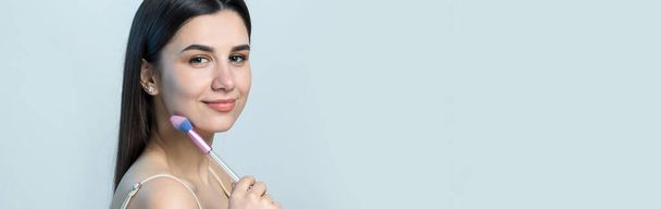 Close-up van een jong meisje in een lichte top op een witte achtergrond die een gezichtsmake-up maakt. Een mooie vrouw houdt een cosmetische borstel bij haar gezicht en glimlacht. - Foto, afbeelding