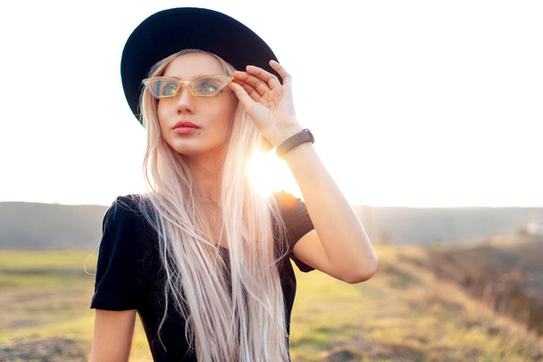 若い美しいブロンドの女の子の肖像画,日没の背景に黒い帽子と黄色の眼鏡をかけて. - 写真・画像