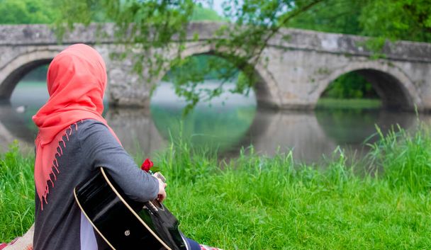 イスラム教徒の女の子のギターを演奏し、橋に向かっている。霞の間川の岸に座ってリラックスしながら - 写真・画像
