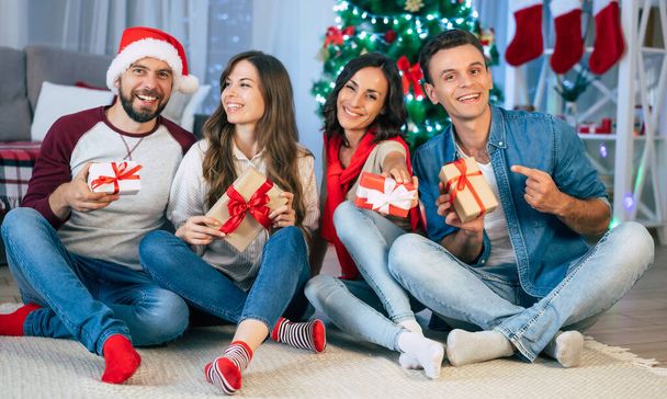 Group of happy and excited best friends are celebrating Christmas and New Year with many gift boxes while sitting on the floor and giving presents to each other on Christmas tree background  - Φωτογραφία, εικόνα