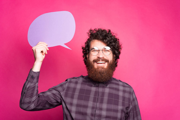 Photo of excited bearded hipster man holding empty speech bubble over pink background and smiling - Photo, Image