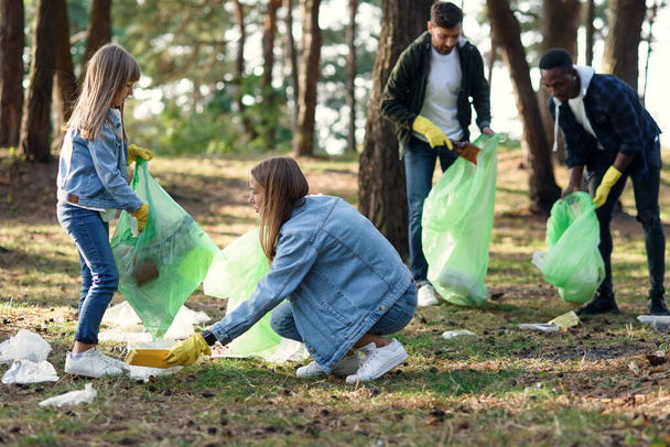 Pretty girl picking up rubbish at the backdrop of multi aged volunteers at the forest. Against pollution concept. - Foto, Imagem
