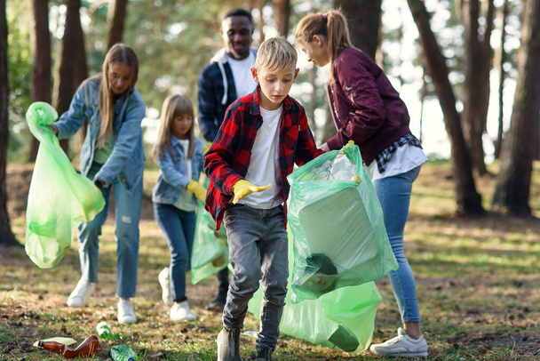 Persistent boy picking up rubbish at the backdrop of multi aged volunteers at the park. Against pollution concept. - Foto, imagen