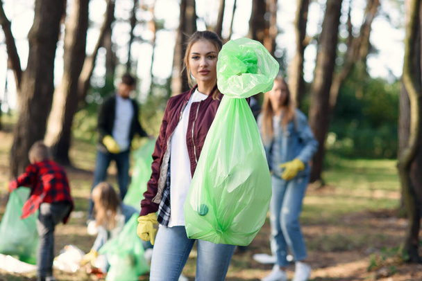 Pretty young woman shows a full rubbish pack in background of his friends volunteers picking up rubbish at park. - Fotografie, Obrázek