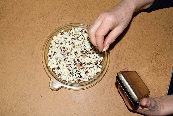 Woman eating popcorn sits at the table with the phone. - Photo, Image