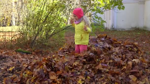 gek kind meisje springen op kleurrijke herfst bladeren stapel in huis achtertuin - Video