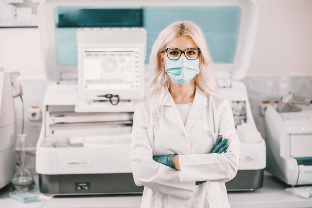 Atrakcyjna blond asystentka laboratoryjna w sterylnym białym mundurze, z maską na twarzy i gumowymi rękawiczkami stojącymi w laboratorium z krzyżowanymi ramionami. - Zdjęcie, obraz