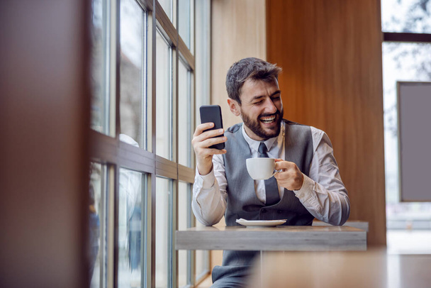 Young attractive bearded employee sitting in coffee shop holding cup of coffee and reading funny message from colleague. - 写真・画像