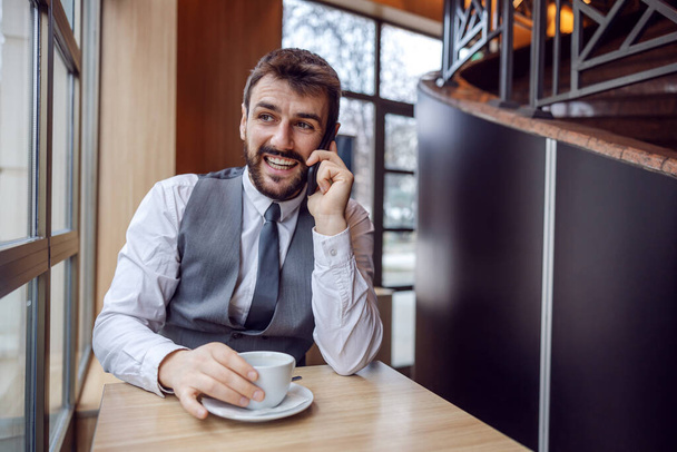 Young smiling businessman sitting in coffee shop on a coffee break and having phone call from important client. - 写真・画像