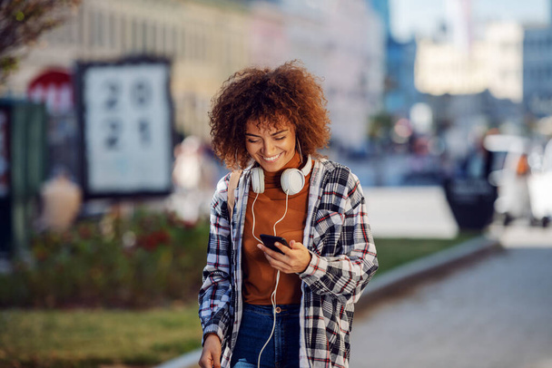 Junge attraktive gemischte Rasse Mädchen mit lockigem Haar gekleidet lässig zu Fuß auf der Straße und mit Smartphone. - Foto, Bild