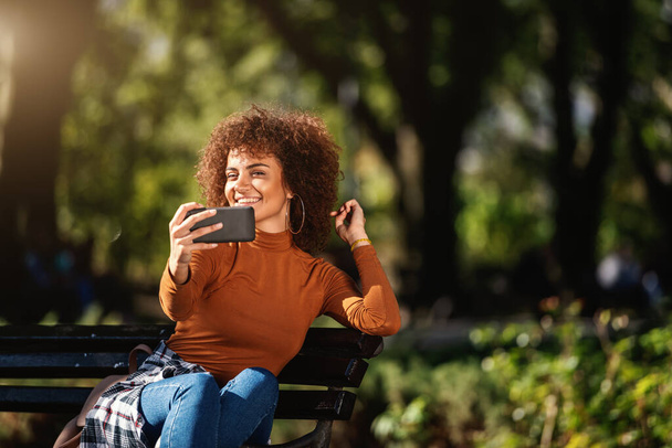 Houkutteleva sekarotuinen tyttö istuu penkillä puistossa ja ottaa selfie. Hän julkaisee sen sosiaalisessa mediassa.. - Valokuva, kuva