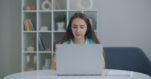 diák nő ül asztalnál otthon viselni headset beszélni távolról videohívás app és laptop eszköz e-learning nyelvet tanul e tutor vagy osztálytársa nem közös feladat távolról, modern tech koncepció - Felvétel, videó