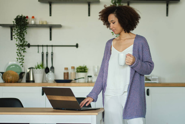 Happy woman holding a cup of coffee and checks mail on his laptop in her kitchen. Young african american female drinking coffee in her kitchen. relaxed morning concept - Valokuva, kuva