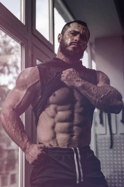 strong young caucasian man athlete with beard showing his perfect physique abdominal muscle near window in sport gym - Zdjęcie, obraz