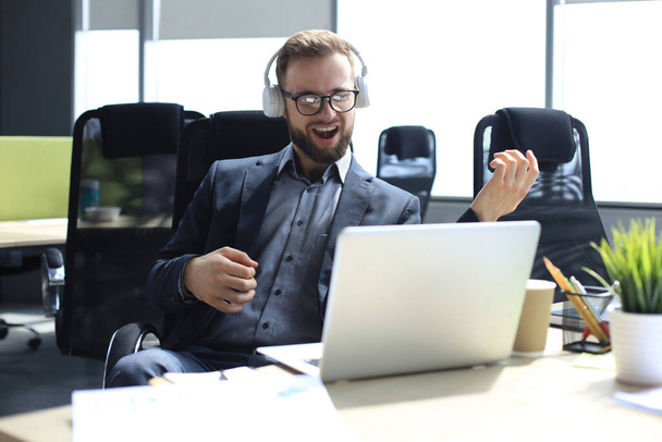 Smiling handsome businessman relaxing and listening music in earphones in a modern office - Photo, Image
