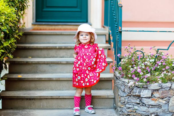 Portrét krásné malé nádherný batole dívka v růžové letní vzhled oblečení, módní šaty, kolena ponožky a klobouk. Šťastné zdravé dítě pózuje před barevným domem. - Fotografie, Obrázek