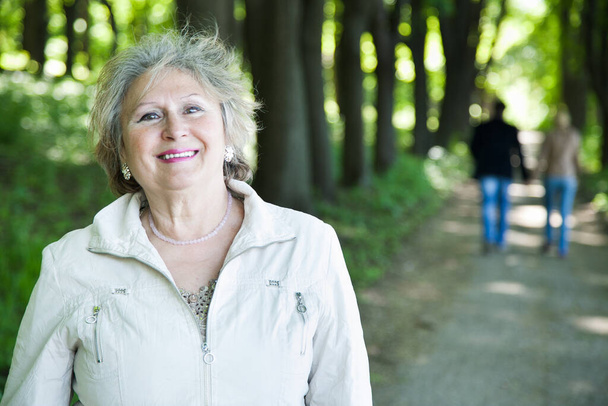 smiling senior aged woman outdoors with blurred walking couple  - Photo, Image