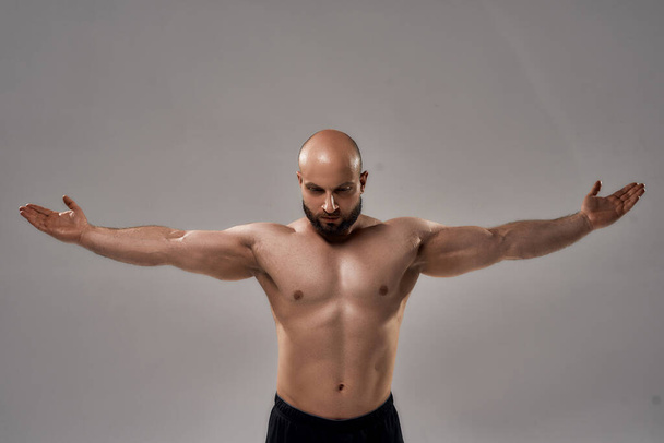 Showing his perfect body. Muscular strong bald man with naked torso standing with outstretched arms isolated over grey background - Fotografie, Obrázek