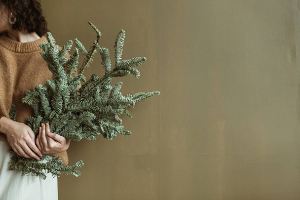 Süveter ve etekli genç bir kadın zeytin duvarına köknar dalları tutuyor. Minimalist moda festivali Noel / Yeni Yıl konsepti. - Fotoğraf, Görsel