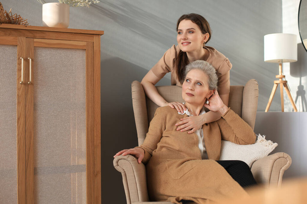 Пожилая средняя мать сидит в кресле, а ее дочь обнимается, смотрит в сторону, доверенные отношения - Фото, изображение
