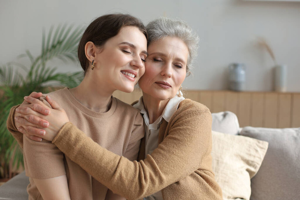Cheerful young woman is embracing her middle aged mother with closed eyes hugging, touching cheeks, sitting on couch at home. Happy trusted relations. Family concept - Photo, Image
