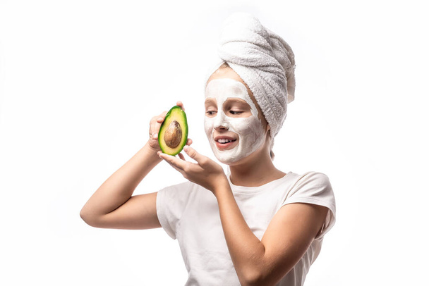 Vtipná holčička s obličejovou maskou, drží avokádové ovoce. Dospívající dívka se stará o svou kůži, čistí póry. Maska obličeje v lázních, koncepce péče o pleť. - Fotografie, Obrázek