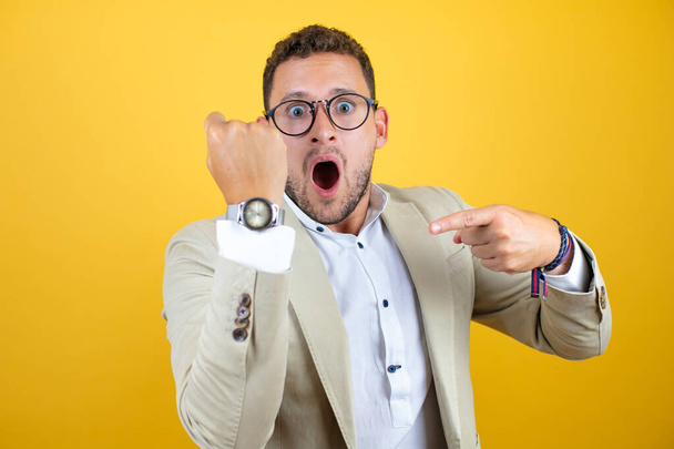 Young handsome businessman wearing suit over isolated yellow background surprised and pointing his watch because it's late - Фото, изображение