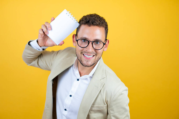 Jeune homme d'affaires beau en costume sur fond jaune isolé souriant et montrant carnet vierge dans sa main - Photo, image