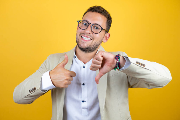 Young handsome businessman wearing suit over isolated yellow background Doing thumbs up and down, disagreement and agreement expression. Crazy conflict - 写真・画像