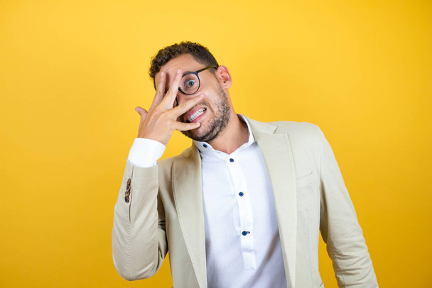 Young handsome businessman wearing suit over isolated yellow background peeking in shock covering face and eyes with hand, looking through fingers with embarrassed expression - 写真・画像