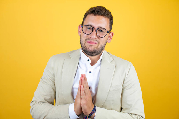 Mladý pohledný podnikatel v obleku přes izolované žluté pozadí žebrání a modlení s rukama dohromady s výrazem naděje na tváři velmi emocionální a ustaraný - Fotografie, Obrázek