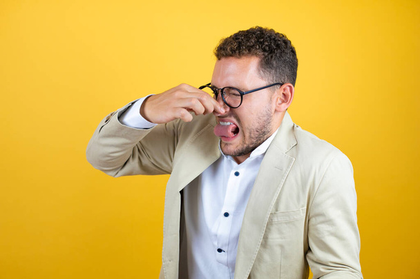 Young handsome businessman wearing suit over isolated yellow background smelling something stinky and disgusting, intolerable smell, holding breath with fingers on nose - Photo, Image