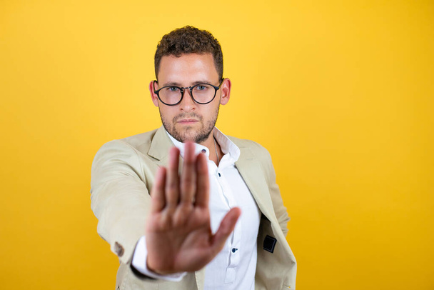 Young handsome businessman wearing suit over isolated yellow background serious and doing stop sing with palm of the hand. - Foto, imagen