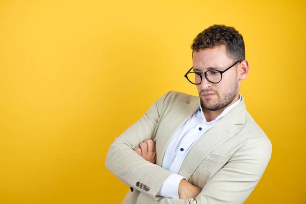 Молодой красивый бизнесмен в костюме на изолированном желтом фоне скептик и нервный, неодобрительное выражение лица со скрещенными руками - Фото, изображение