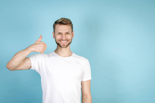 Good deal concept. Successful and confident adult man in white t-shirt showing thumb up, smiling wide, looking at camera, standing isolated on blue copy space background - Photo, Image