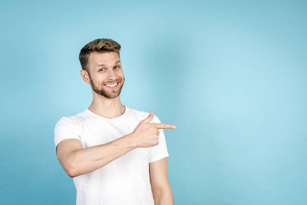 Щасливий і усміхнений молодий чоловік у білій футболці, що вказує пальцем убік, дивиться на камеру, рекомендує новини, стоїть ізольовано на синьому фоні копіювання. Продаж та реклама зі знижкою
 - Фото, зображення