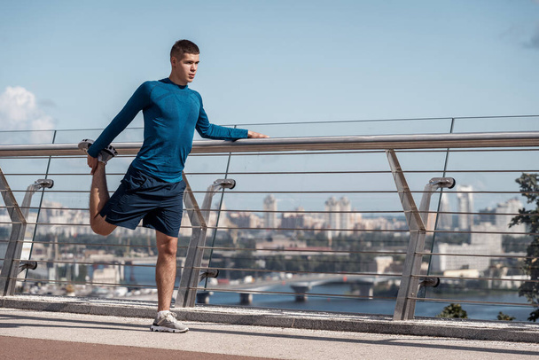 Full length and side view of focused adult sporty guy making sport training outdoors alone, doing exercise, stretching legs before running in city - Φωτογραφία, εικόνα